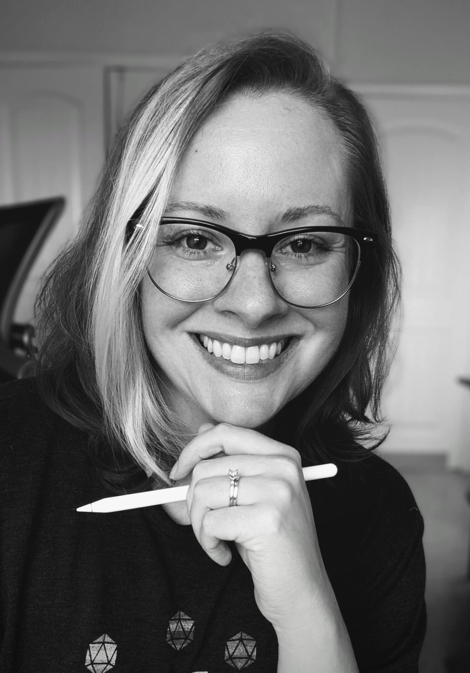 black and white headshot of artist - a pale skinned person with shoulder length hair, wearing black rimmed glasses, holding an Apple pencil in left hand